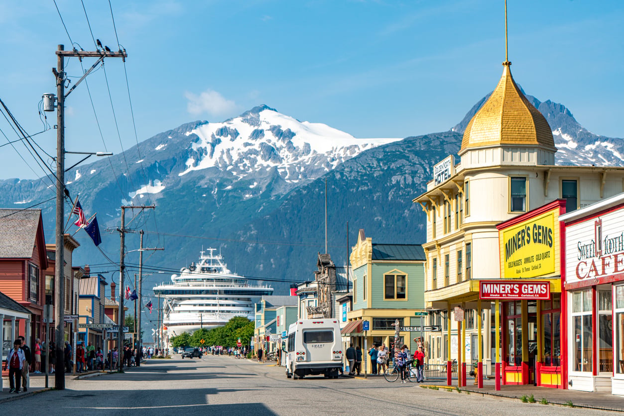 A busy street in a small town with colorful buildings, a cruise ship docked nearby, people walking, and snow-capped mountains in the background under a clear blue sky—one of those nostalgic travel destinations that captures the charm of days gone by.