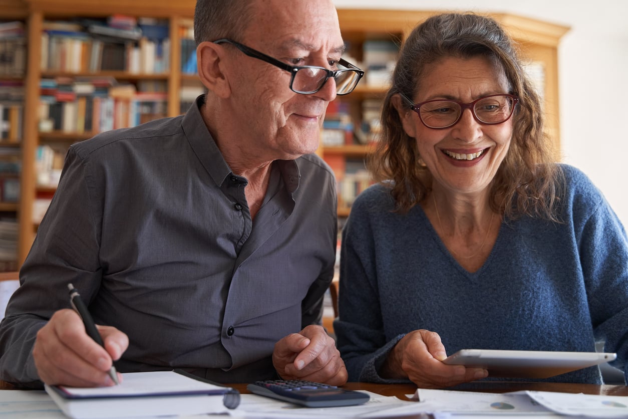 An older man writes in a notebook while an older woman smiles and holds a tablet. They sit together at a table with papers, a calculator, and bookshelves in the background.