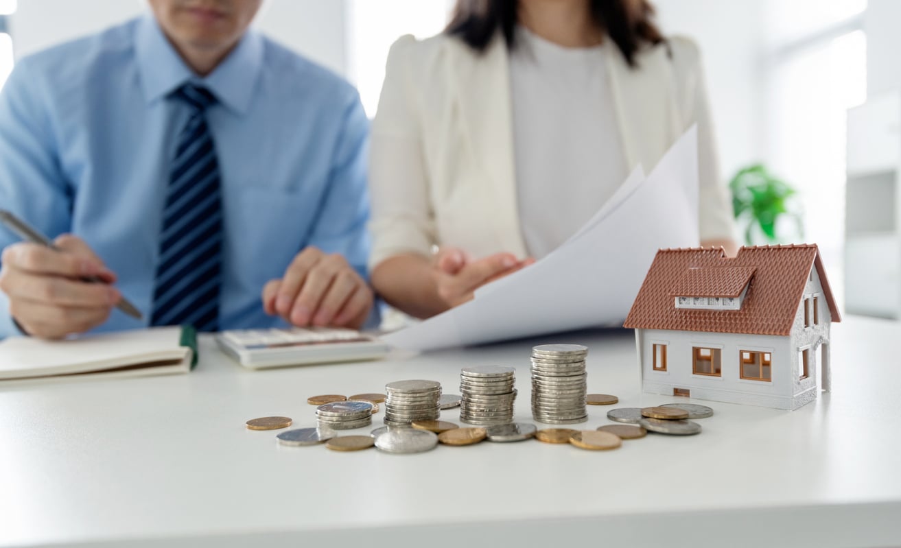Two people reviewing documents at a desk with a calculator, coins, and a small house model, symbolizing financial planning or real estate investment.