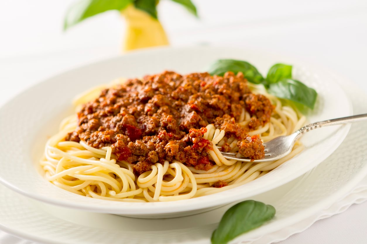 A plate of spaghetti topped with meat sauce, garnished with fresh basil leaves, with a fork twirling some pasta on the side.