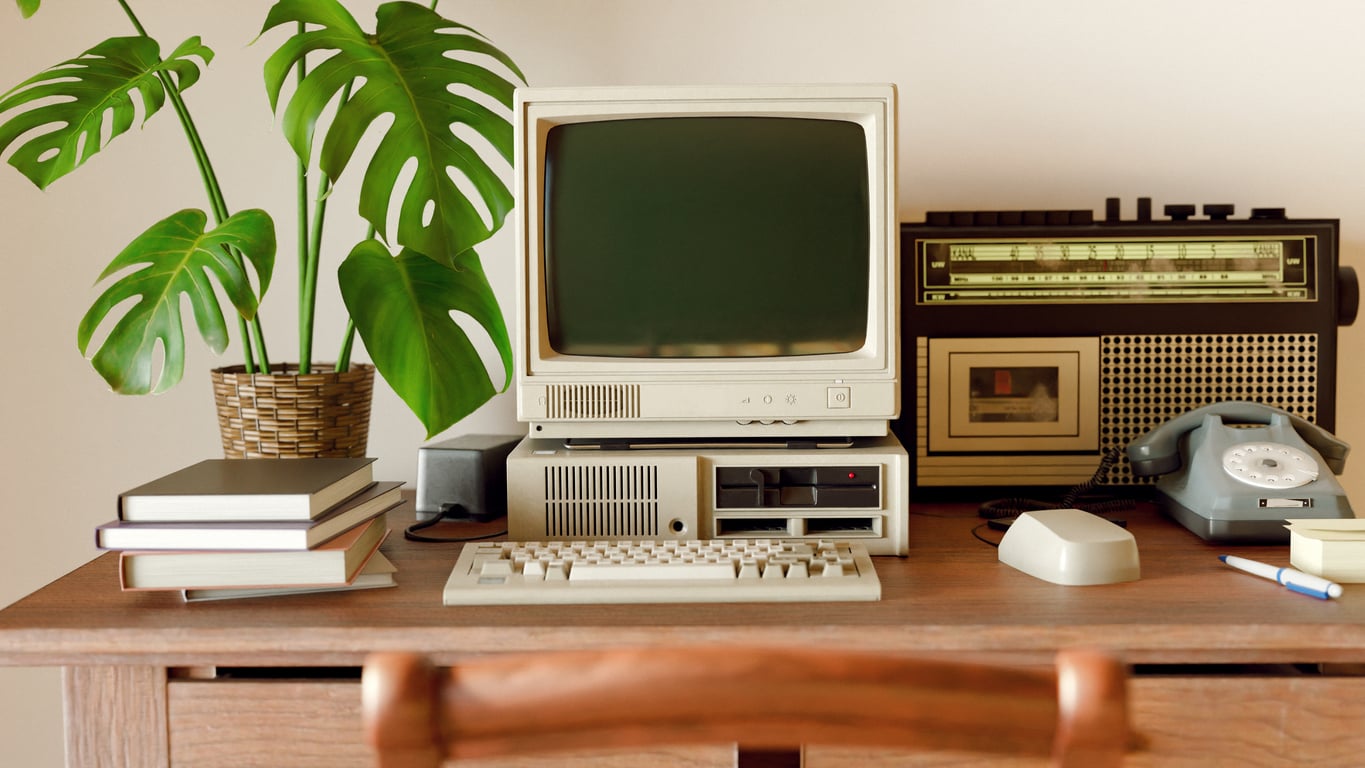 A vintage desk setup with an old computer, monitor, mouse, rotary phone, radio, stack of books, a pen, and a potted plant with large leaves. The scene has a nostalgic, retro feel.