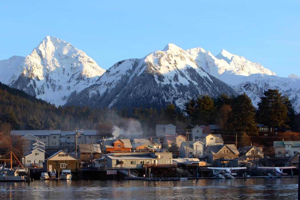 A small town with houses sits by the water, smoke rising from a chimney. Snow-covered mountains tower in the background under a clear blue sky, creating a picturesque scene often found among travel facts about the United States. Pine trees line the edge of the town.