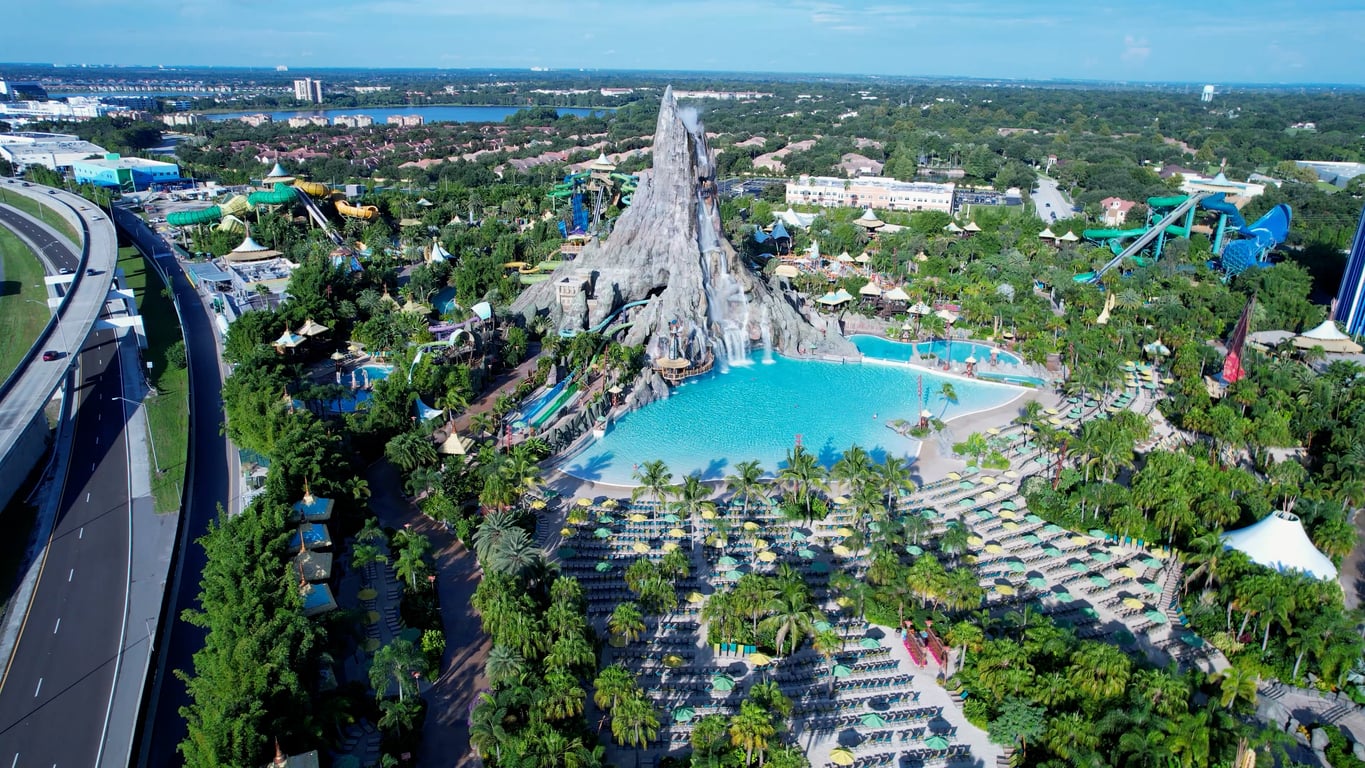 Aerial view of a tropical-themed water park with a large volcano centerpiece, a wave pool, water slides, palm trees, and lounge chairs, surrounded by greenery and nearby roads—despite the stunning setting and fun attractions, some visitors note the presence of bad food options at the park’s eateries.