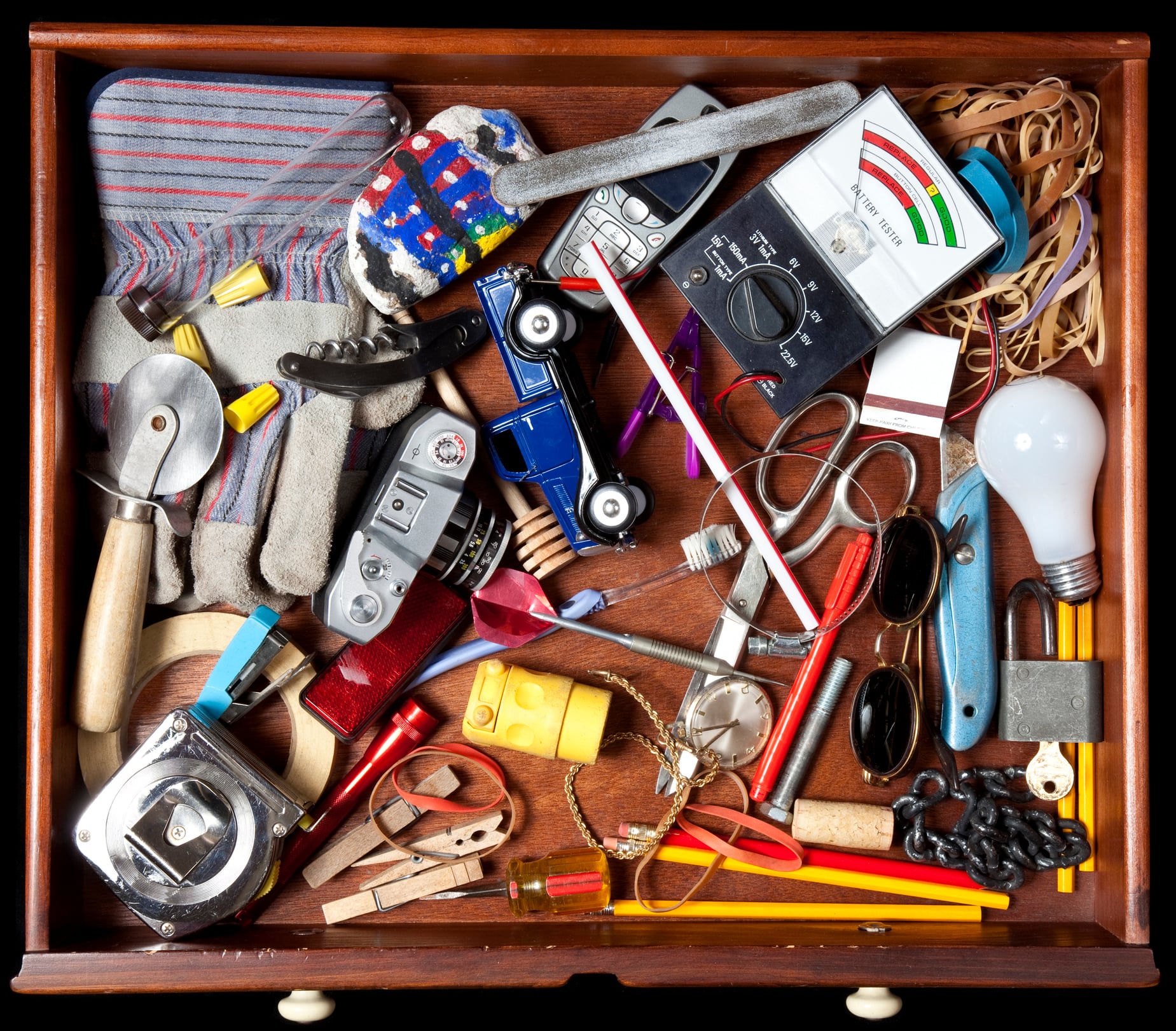 A messy drawer filled with various items, just like you’d find in the 80s, including a lightbulb, toy cars, tape measure, sunglasses, rubber bands, gloves, pencils, remote control, keys, and other small objects.