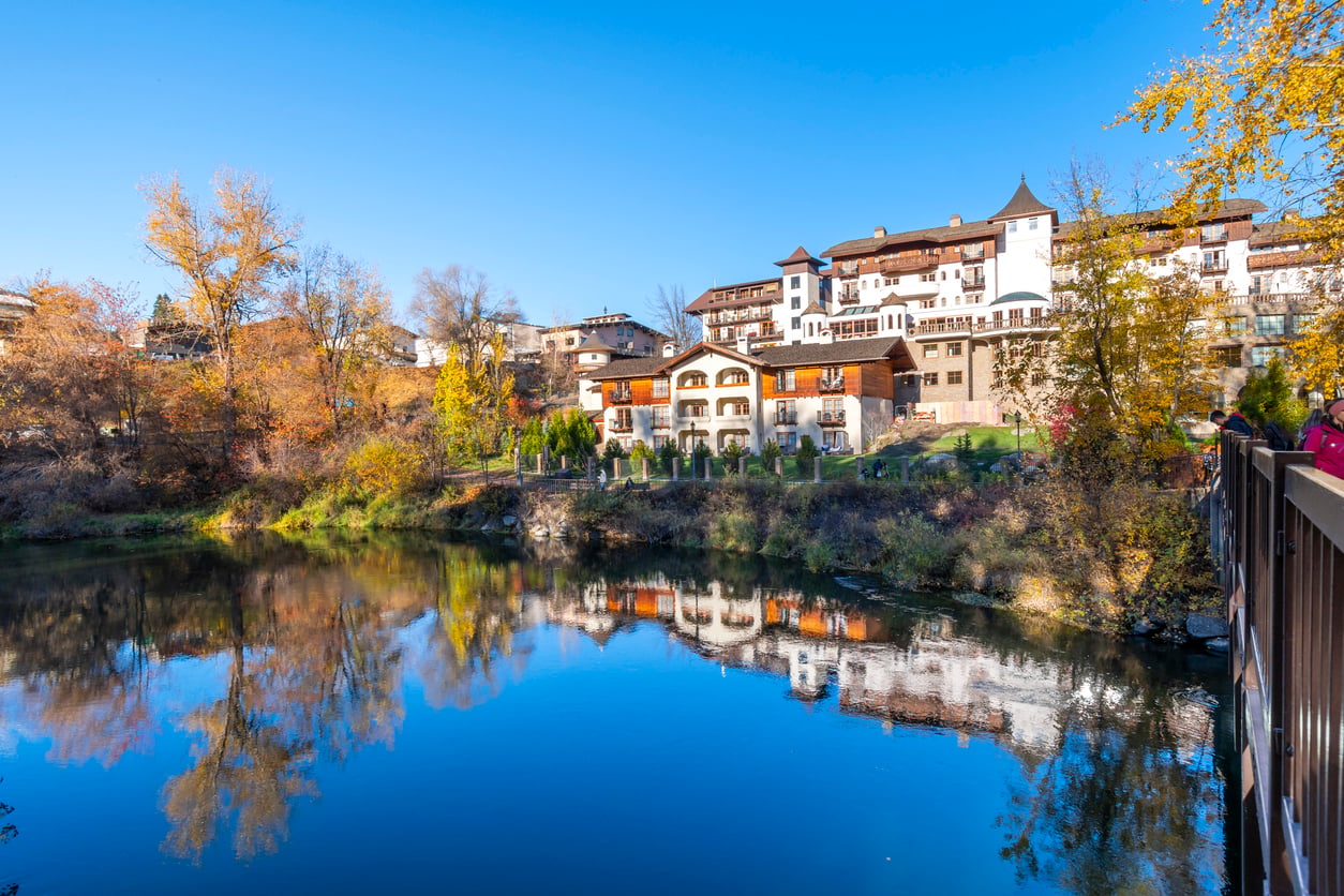 Buildings with European-inspired architecture are reflected in a calm river, surrounded by autumn trees under a clear blue sky—capturing the charm often found in the best fall towns in America. A wooden railing is visible on the right side of the image.