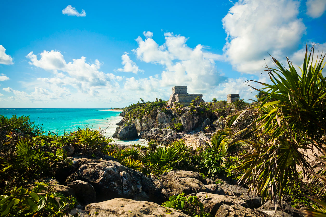 Ancient stone ruins sit atop a cliff overlooking turquoise ocean waters and a sandy beach, surrounded by lush green tropical plants under a bright blue sky with scattered clouds.