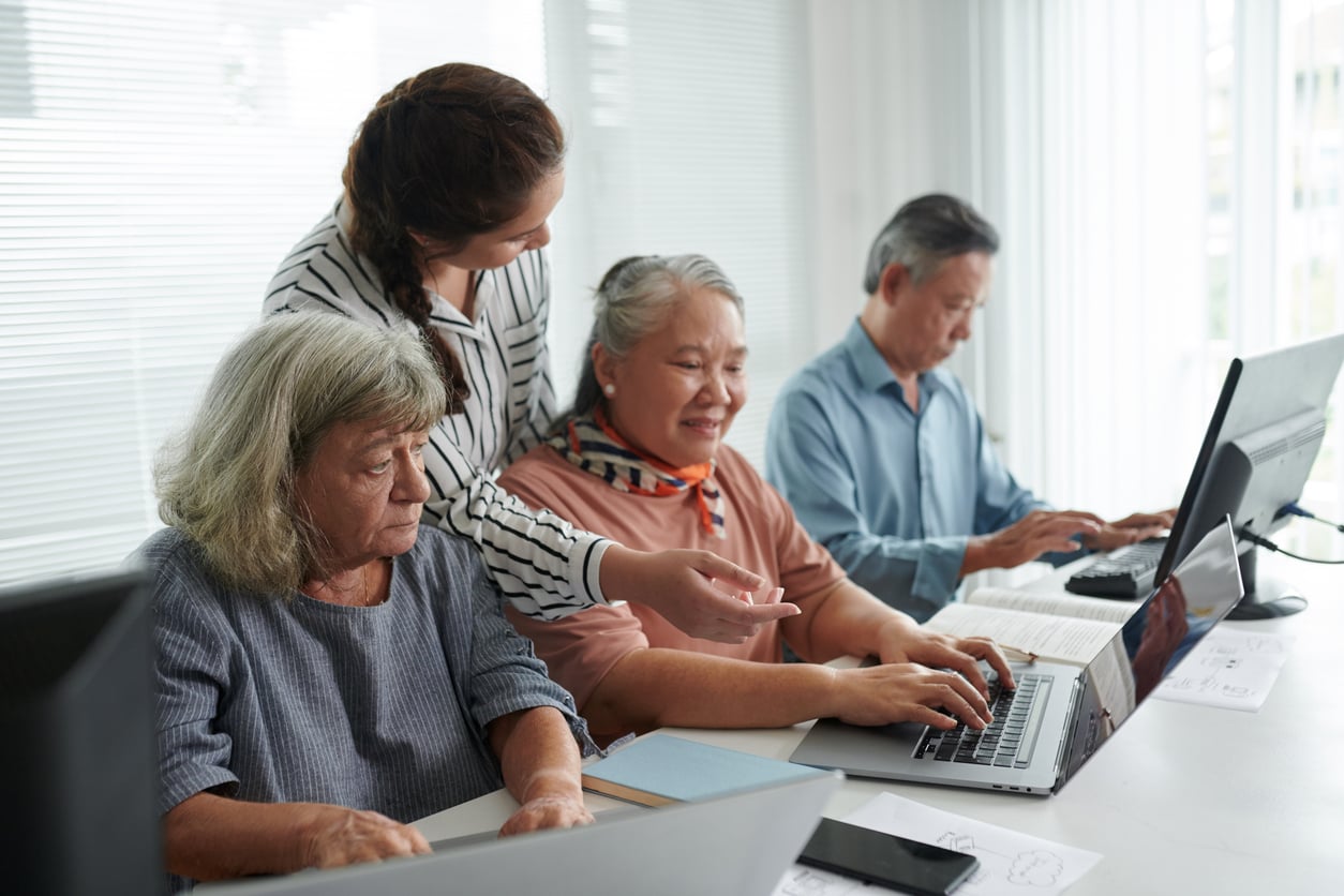 A young woman helps three older adults use laptops in a bright room. The group sits at a table, appearing focused and engaged in learning together, reflecting the positive impact of Baby Boomer changes in embracing new technology.