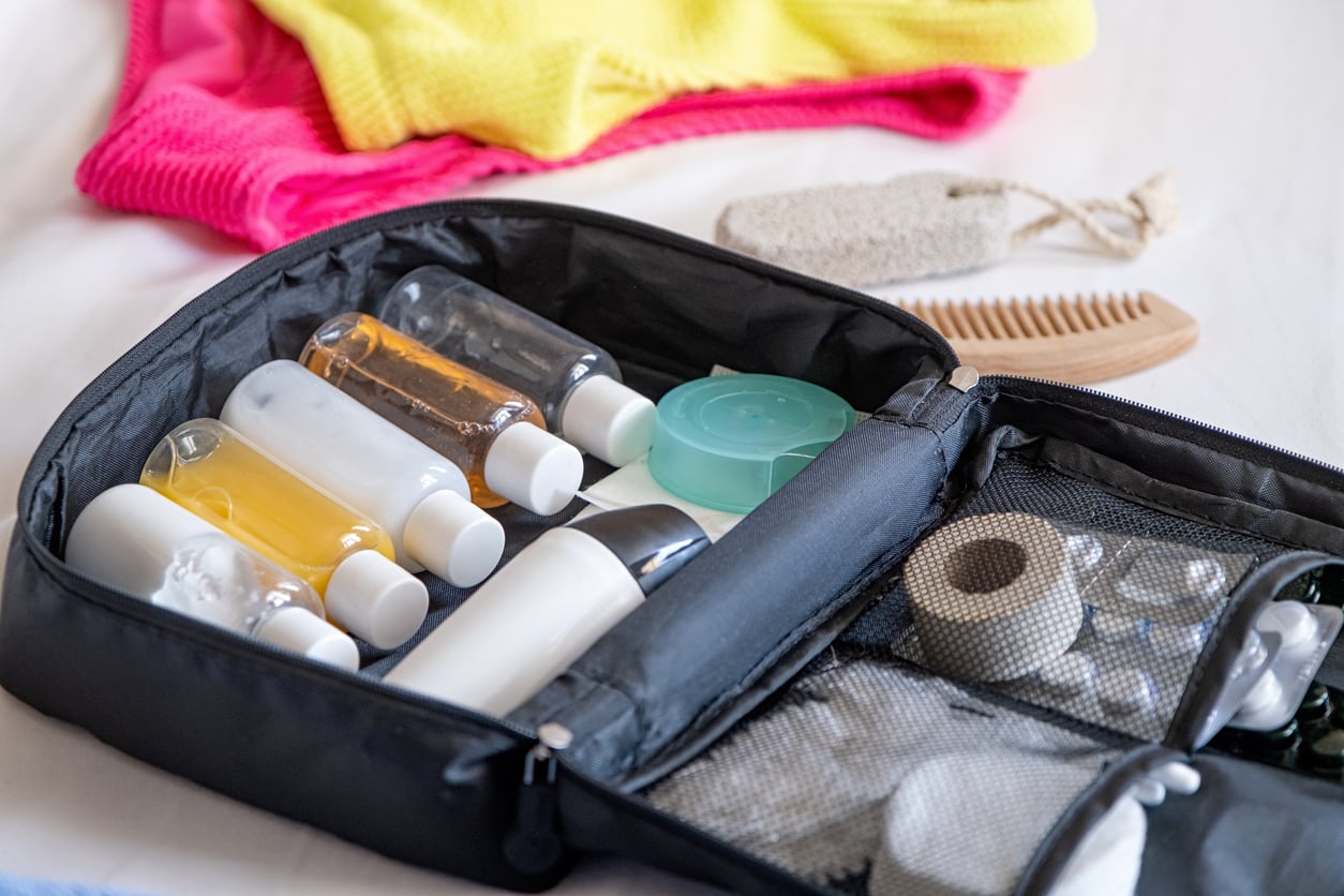 A black toiletry bag filled with small bottles, containers, and travel essentials sits open on a bed next to a yellow towel, pink clothing, a comb, and a pumice stone.
