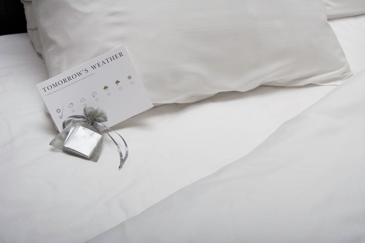 A white bed with pillows, a weather forecast card labeled “Tomorrow’s Weather,” and a small gray drawstring pouch with a silver envelope on the sheets—quiet reminders of the charming things hotels used to offer guests.
