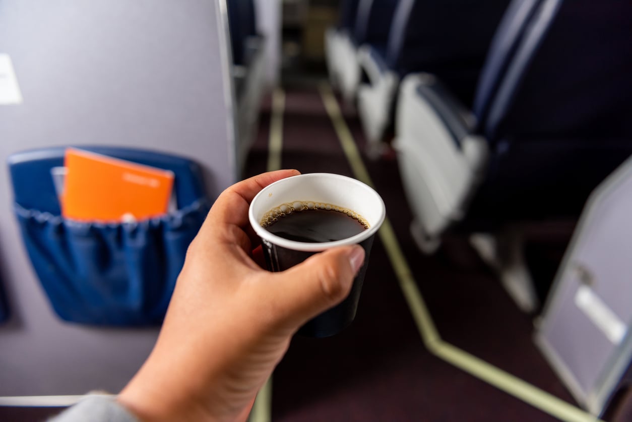 A hand holds a small cup of black coffee inside an airplane cabin, with seats and a pocket holding an orange booklet visible in the background.