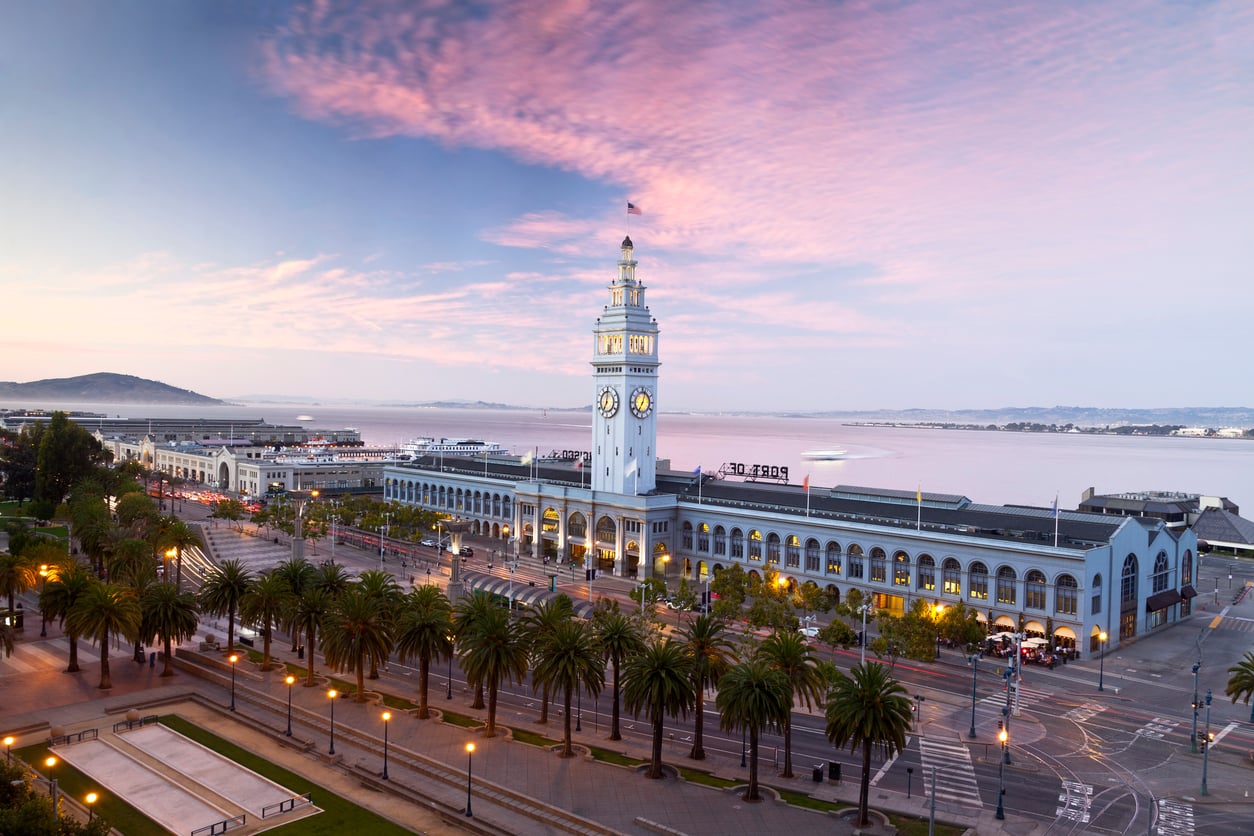 San Francisco Ferry Building at sunrise with clock tower, palm trees, and waterfront; pink and blue sky over calm water in the background—a picturesque scene that stands in stark contrast to tales of bad food sometimes found nearby.