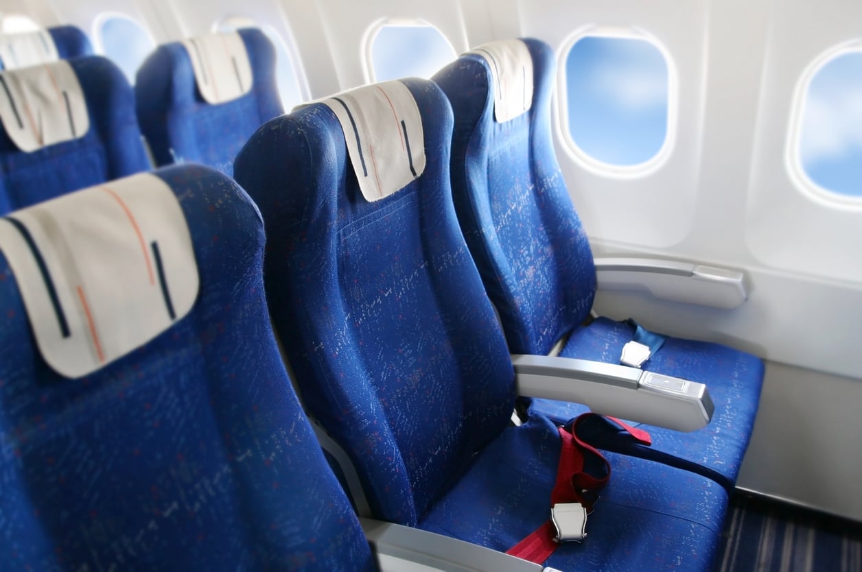 Three empty blue airplane seats by the window, with seat belts fastened and sunlight coming through the windows.