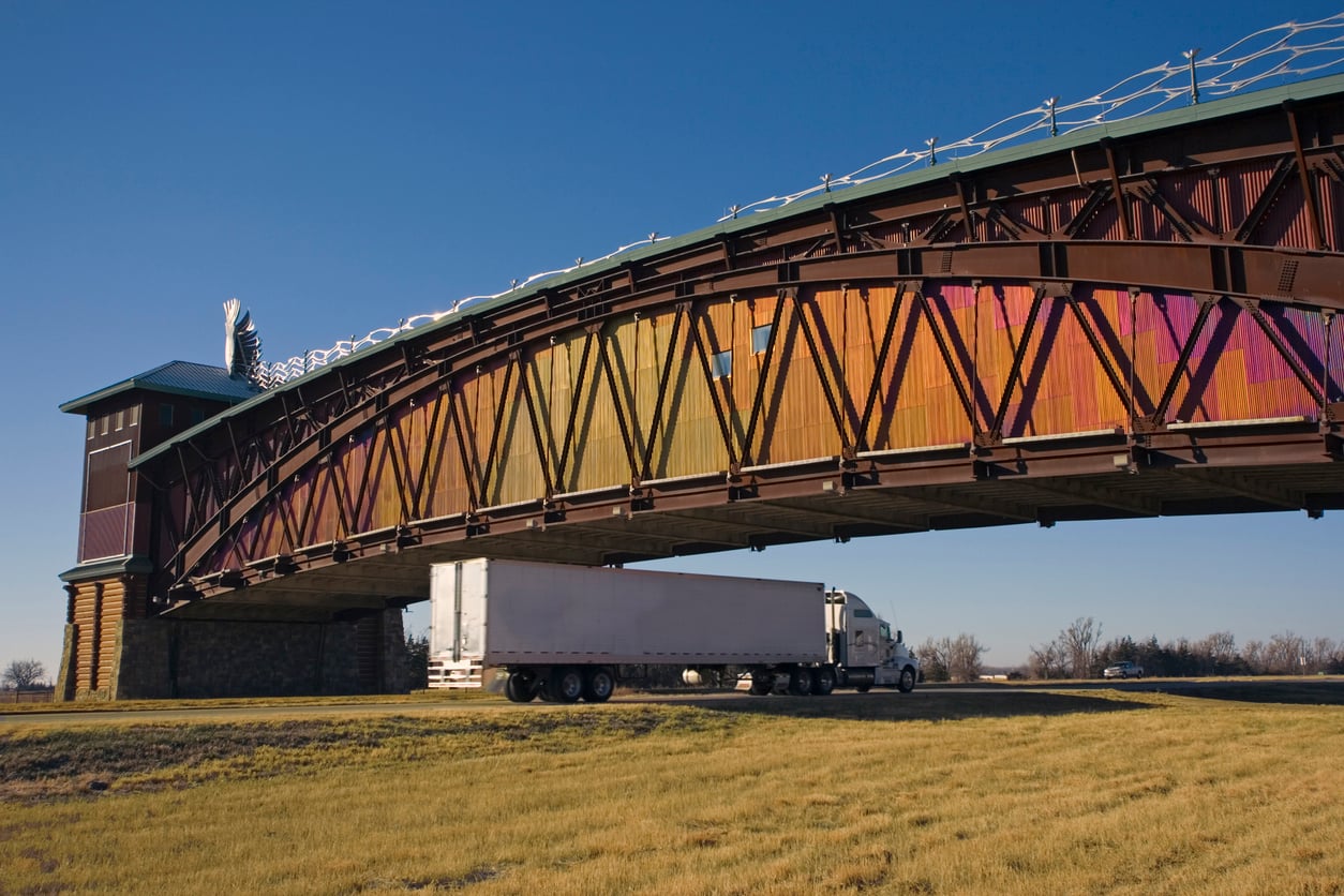 A semi-truck drives under the large, colorful arch of the Great Platte River Road Archway Monument on a clear day with blue sky and grassy ground.