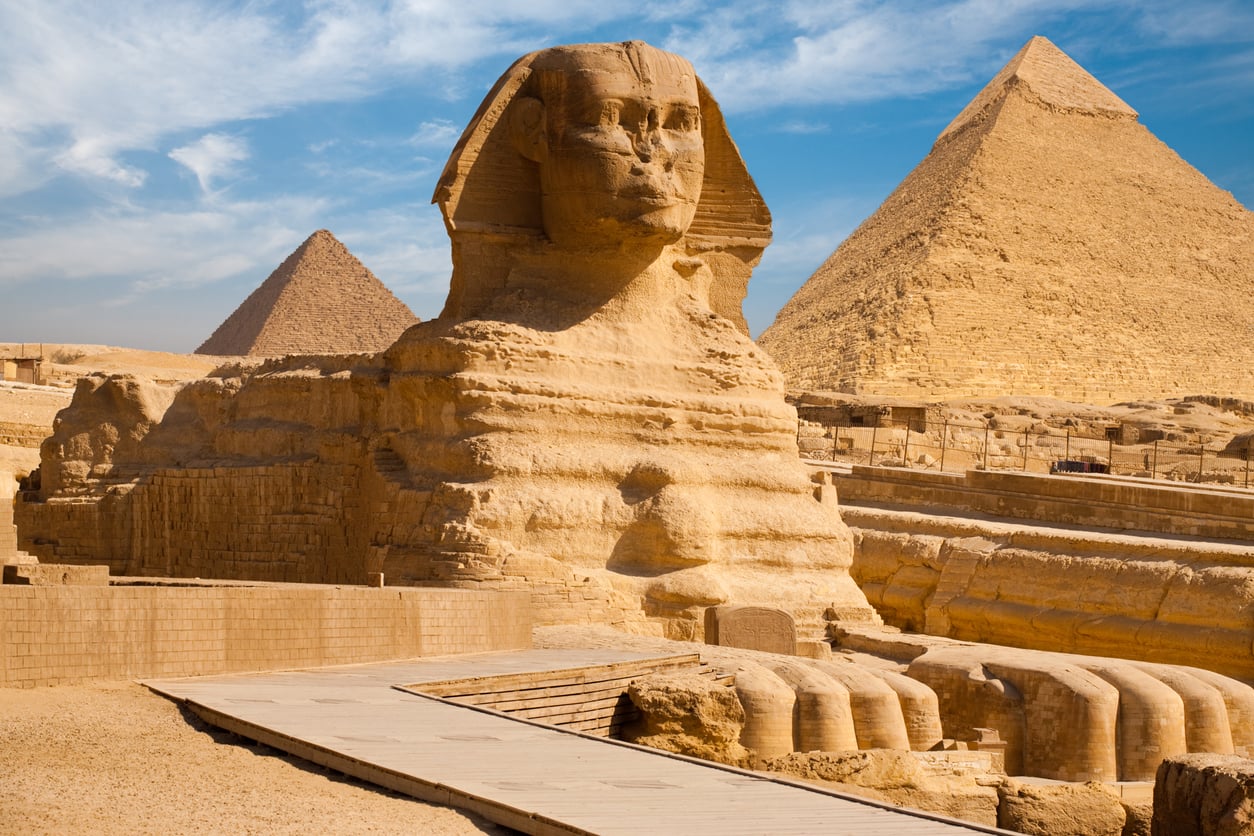 The Great Sphinx of Giza sits in front of two large pyramids under a blue sky with wispy clouds; a sandy stone walkway leads past the monument.