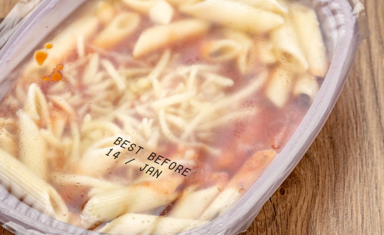 A close-up of a microwave meal with pasta and sauce in a plastic container, covered with condensation. The label reads “BEST BEFORE 14/JAN.” The container rests on a wooden surface.