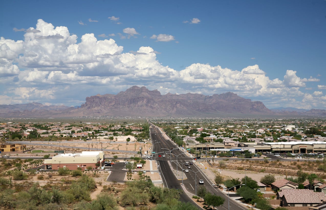 A wide road leads toward distant mountains under a partly cloudy sky, with houses, buildings, and desert vegetation on either side of the road.
