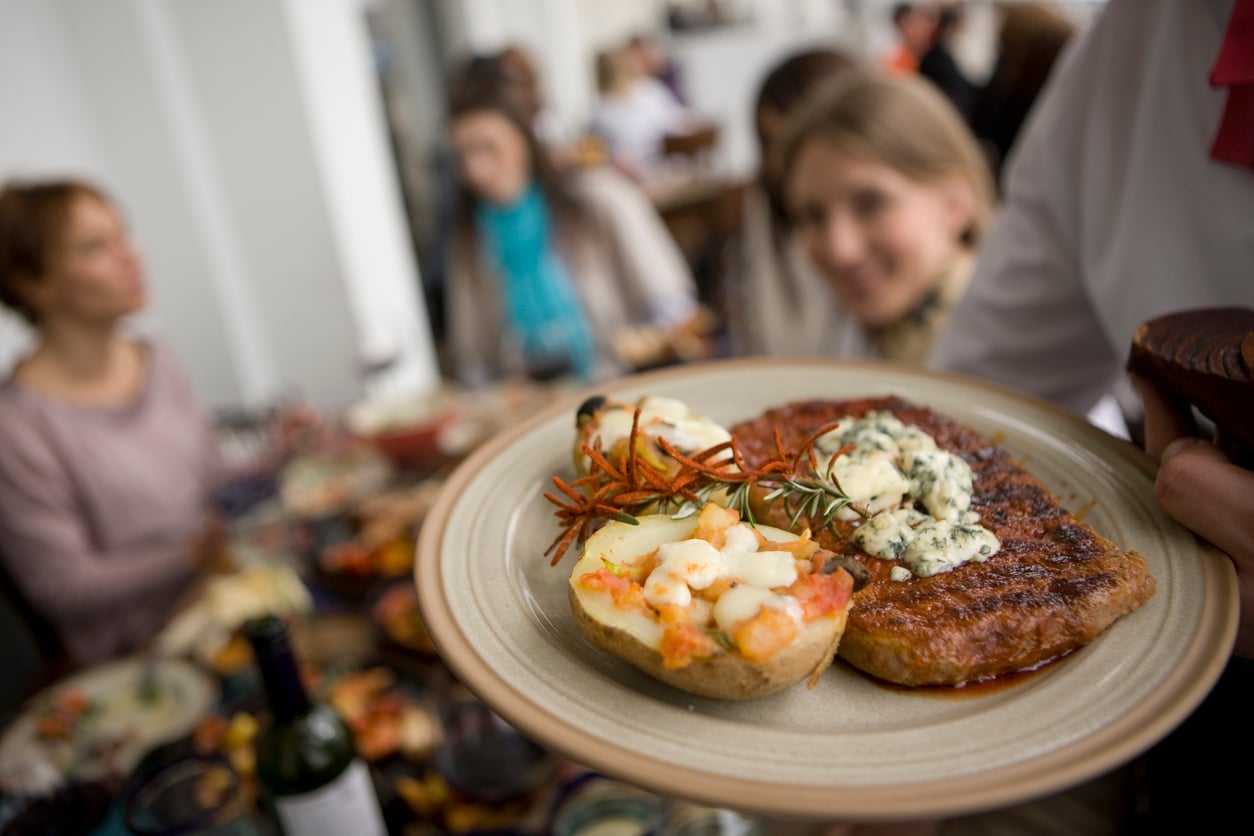 A close-up of a plate with steak topped with blue cheese and herbs, and a baked potato with cheese, held in front of people sitting at a dining table.
