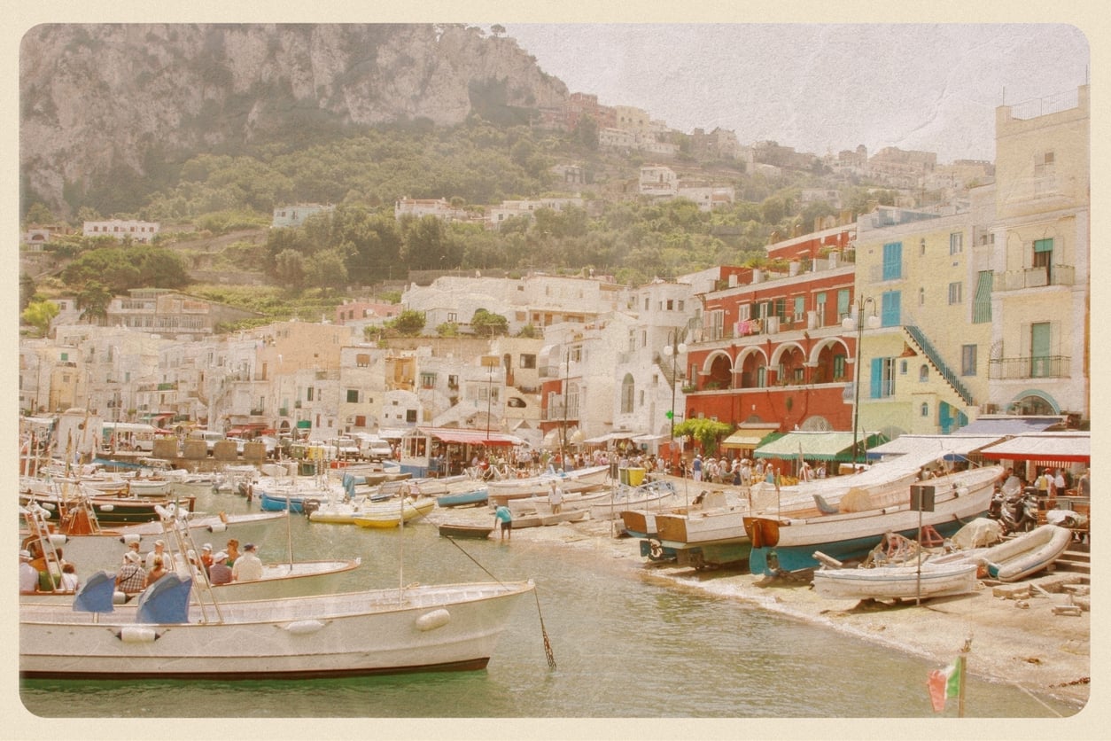 Colorful boats docked by a lively coastal village with pastel buildings and a hillside in the background, under a hazy, vintage filter evoke nostalgia for simpler times—when people strolled near the waterfront and enjoyed charming hospitality reminiscent of things hotels used to offer guests.