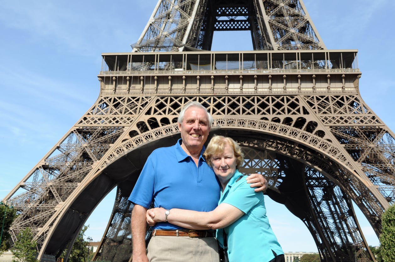 An older couple smiles and hugs in front of the Eiffel Tower on a sunny day, enjoying their visit to Paris—a city that continues to enchant travelers, even as it’s among the travel destinations boomers are skipping in favor of new adventures.