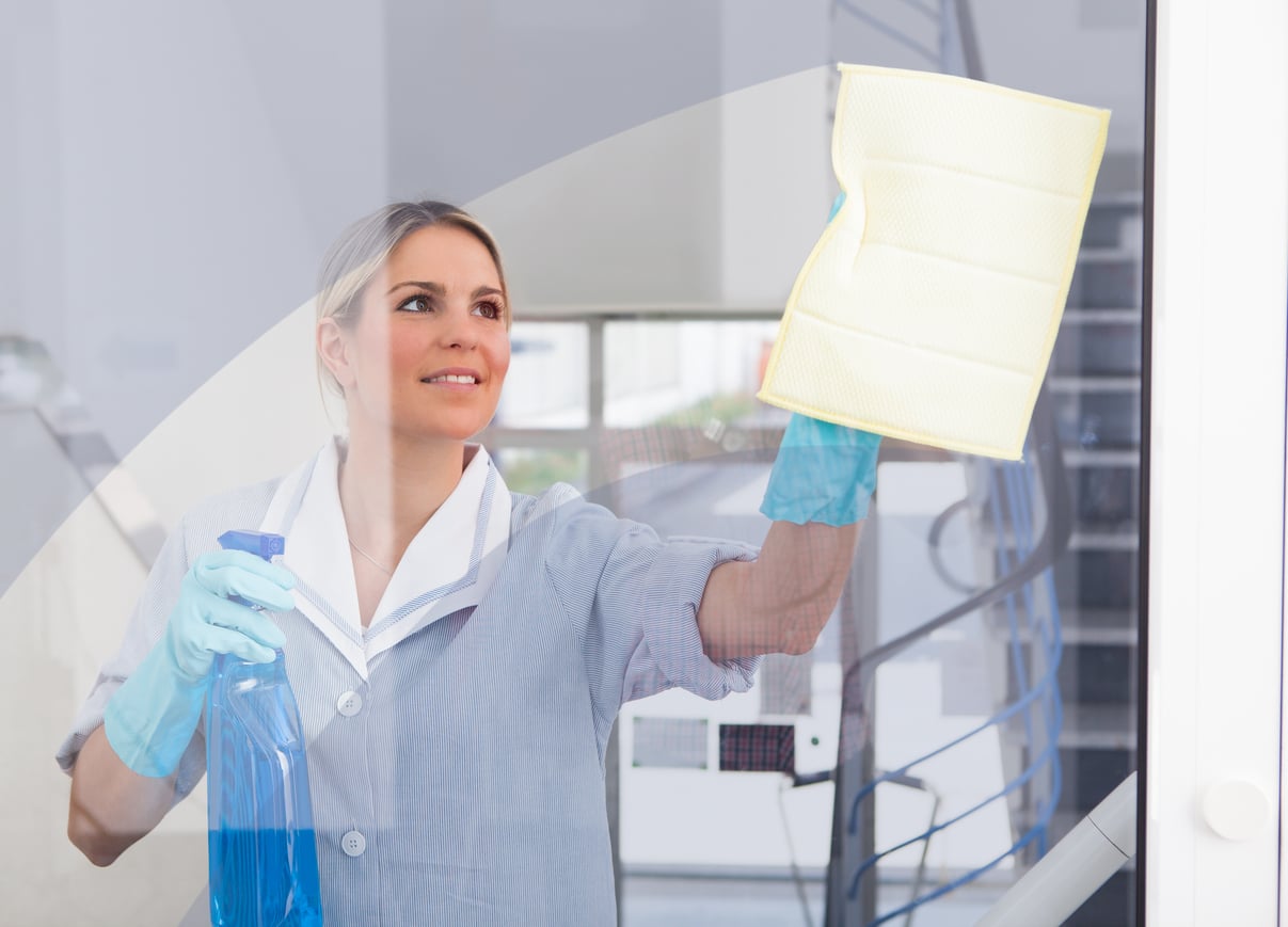 A woman wearing gloves and a cleaning uniform cleans a glass window with a yellow cloth and a spray bottle, smiling as she works indoors near a staircase.