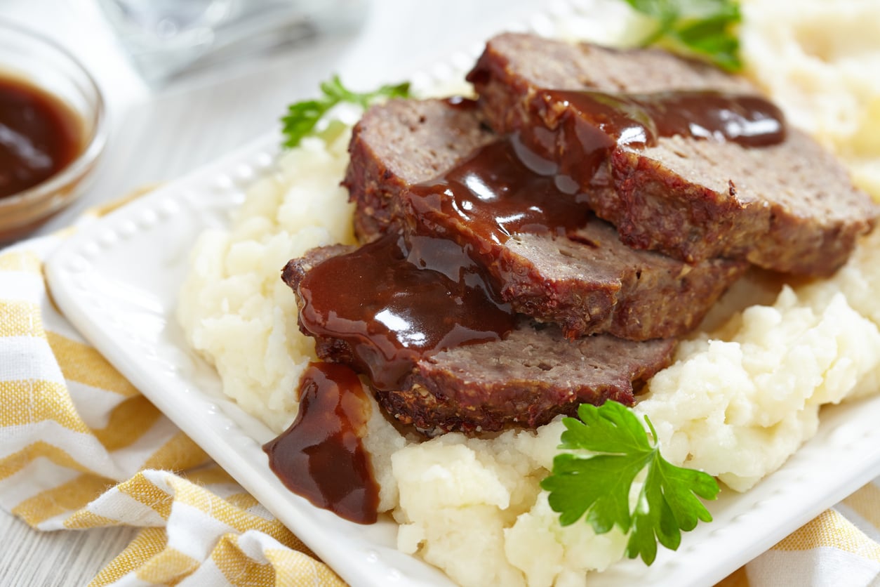 Three slices of meatloaf topped with brown gravy sit on a bed of mashed potatoes, garnished with parsley, on a white rectangular plate—a classic example of the comforting meals middle-class moms made.