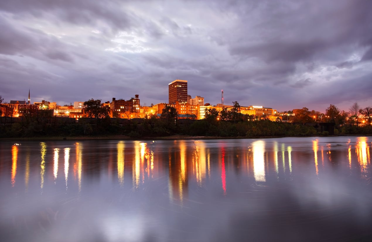 A city skyline at dusk with lit buildings reflecting on a calm river under a cloudy sky.