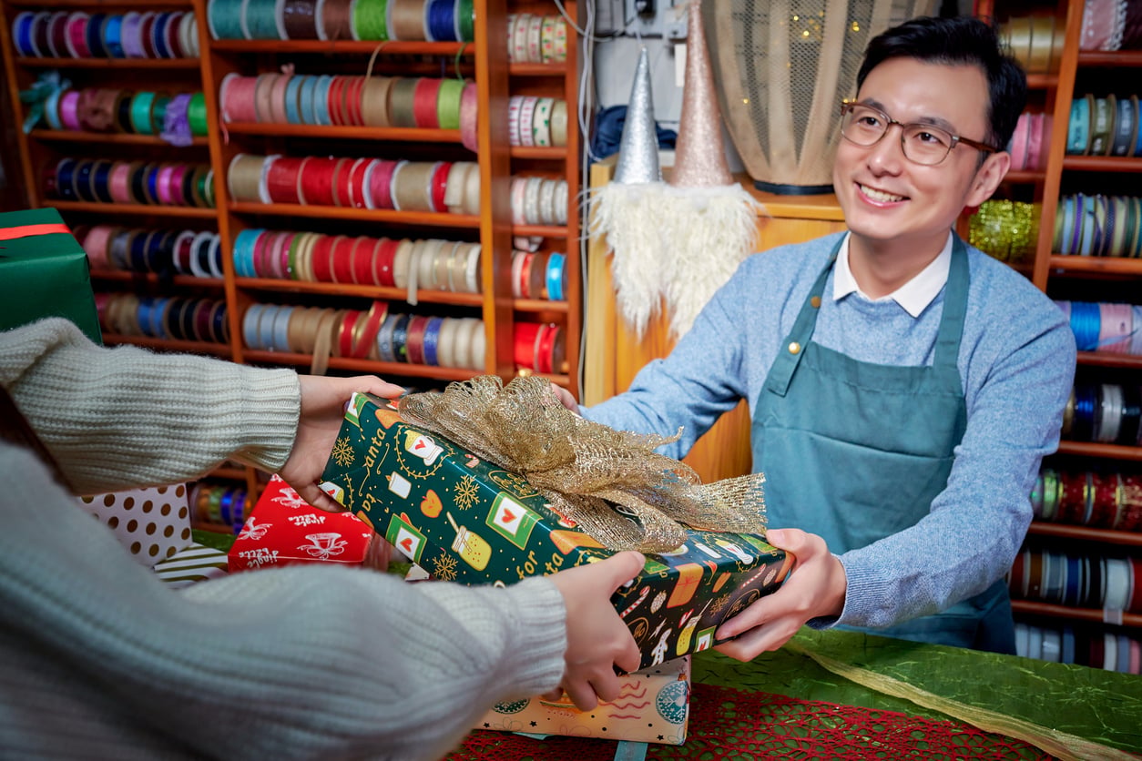 A smiling shop worker in an apron hands a wrapped gift with a gold bow to a customer. Shelves filled with colorful ribbons are in the background.