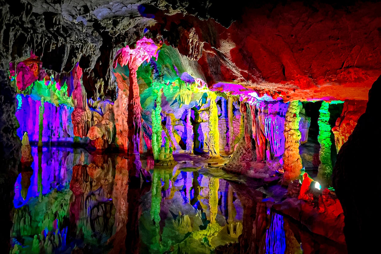 A vibrant cave scene reminiscent of Classic Roadside Attractions, featuring stalactites and stalagmites illuminated by colorful lights in red, green, blue, and purple, reflecting off a calm pool of water below.