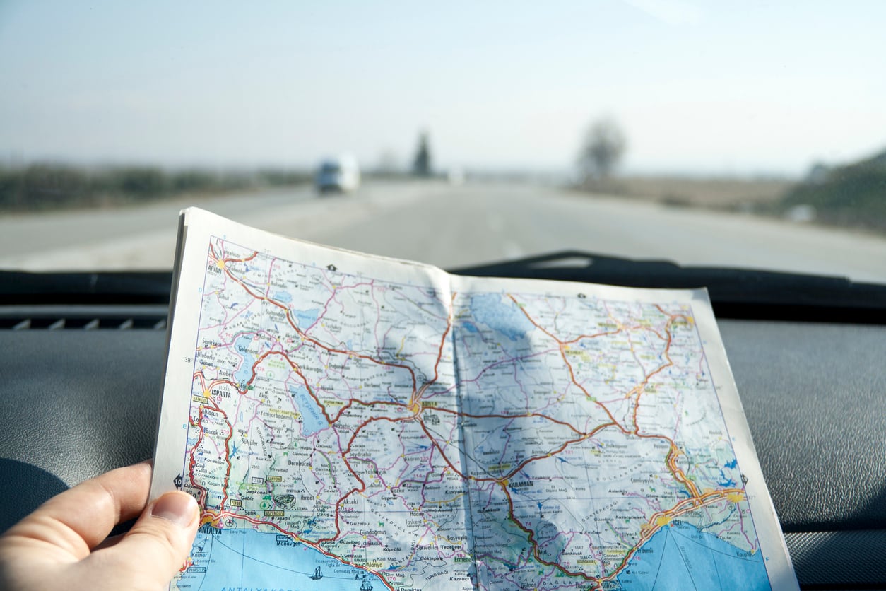 A person holds a road map inside a car, with the map spread open on the dashboard and a highway visible through the windshield on a clear day.