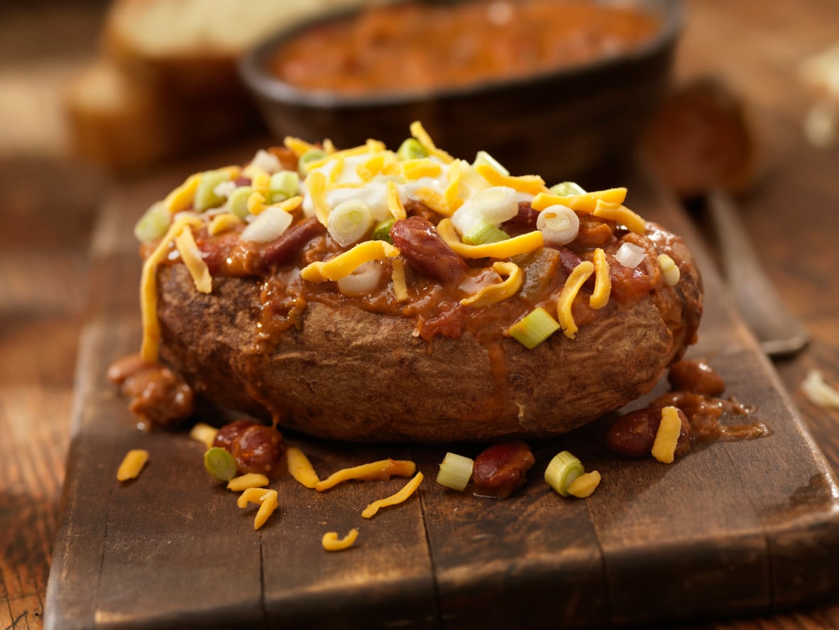 A baked potato topped with chili, melted cheddar cheese, and chopped green onions sits on a wooden board. A bowl of chili and bread are blurred in the background.