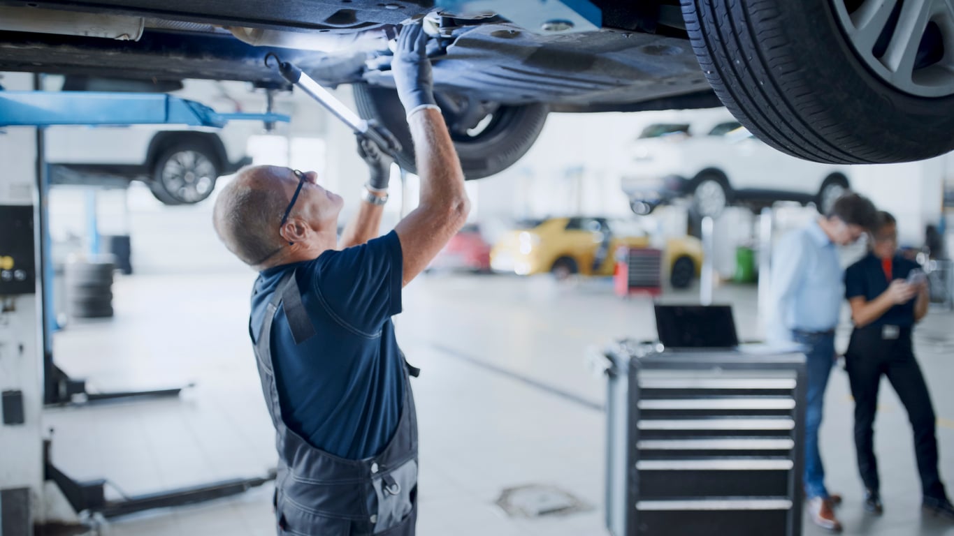 A mechanic in overalls works under a raised car in a garage, using a tool. Other vehicles, a tool chest, and two people in the background are visible in the well-lit workshop.
