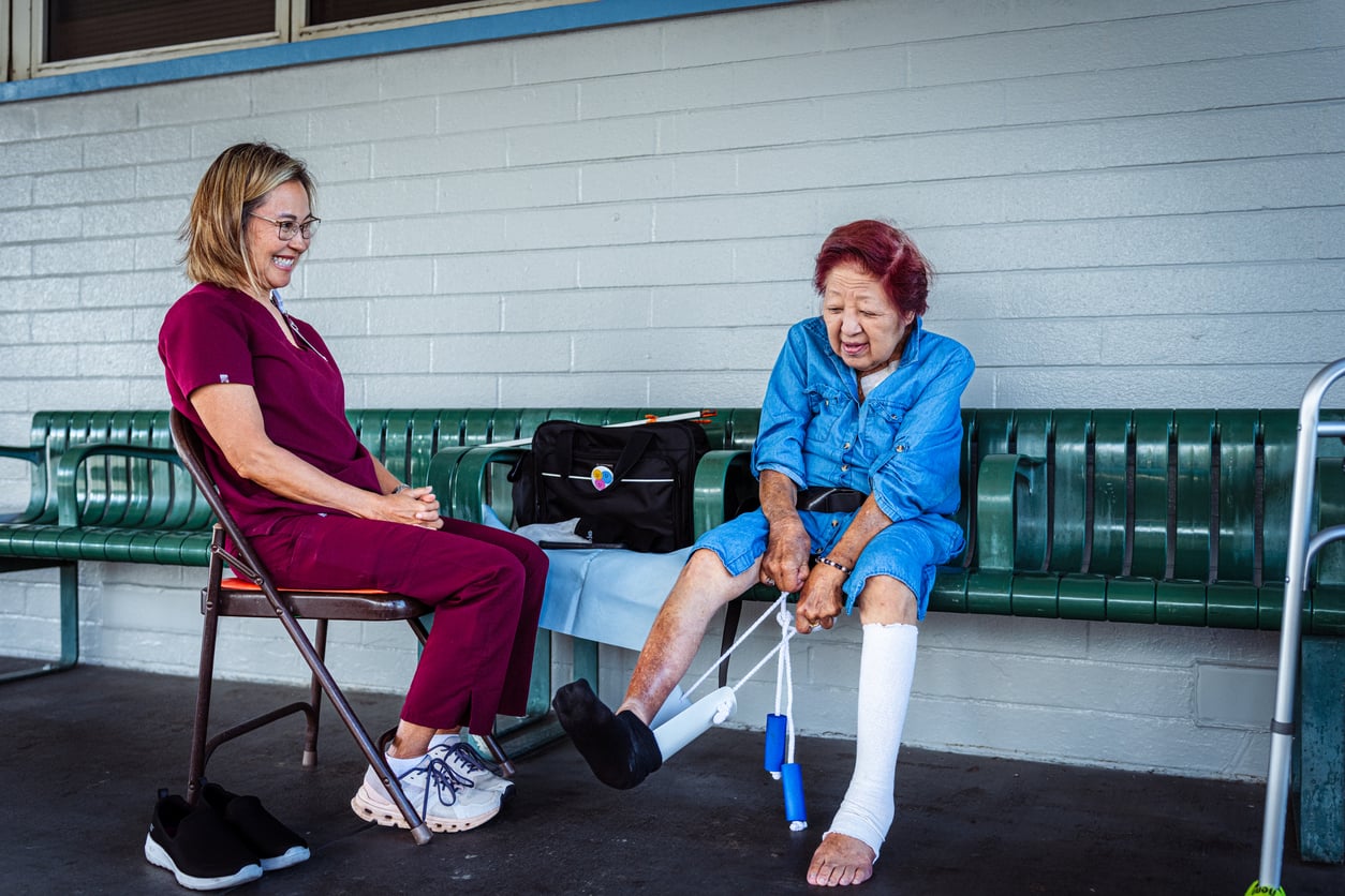 An elderly woman with a bandaged leg uses a dressing aid to put on a shoe, while a healthcare worker in scrubs sits nearby, smiling. They are sitting on a green bench outside, with a bag on the bench.