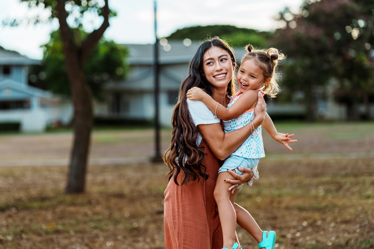 A woman with long hair smiles while holding a laughing young girl in her arms at a park. Trees and houses are visible in the background on a sunny day.
