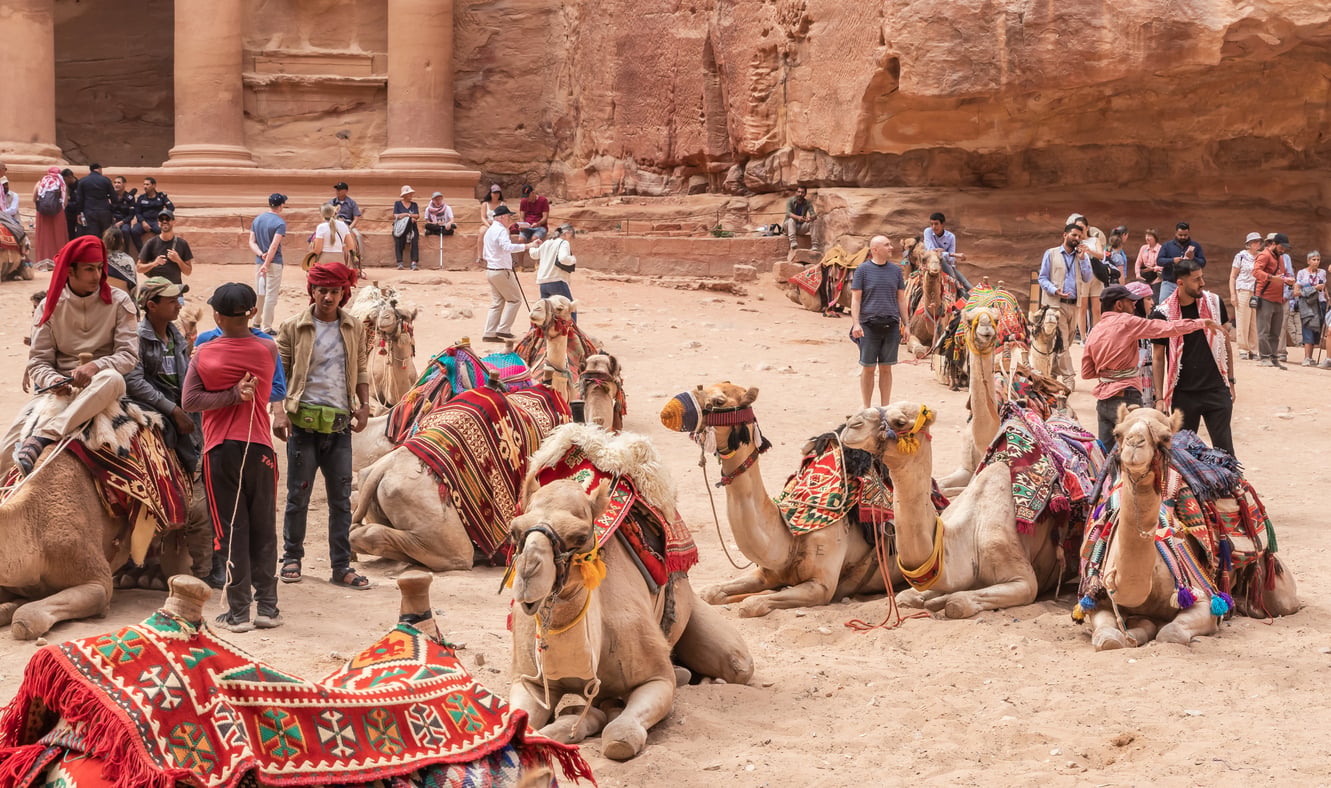 Tourists and locals with camels in colorful saddles gather in front of the ancient stone facade of Petra, Jordan, surrounded by sandy terrain and red rock cliffs, while staying informed about current travel restrictions and advisories to ensure a safe journey.