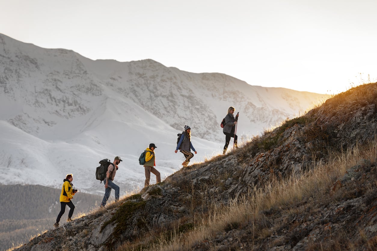 Five hikers with backpacks, showcasing adventurous travel behaviors, walk up a grassy hillside at sunrise, with snow-covered mountains in the background and the sun peeking over the ridge.