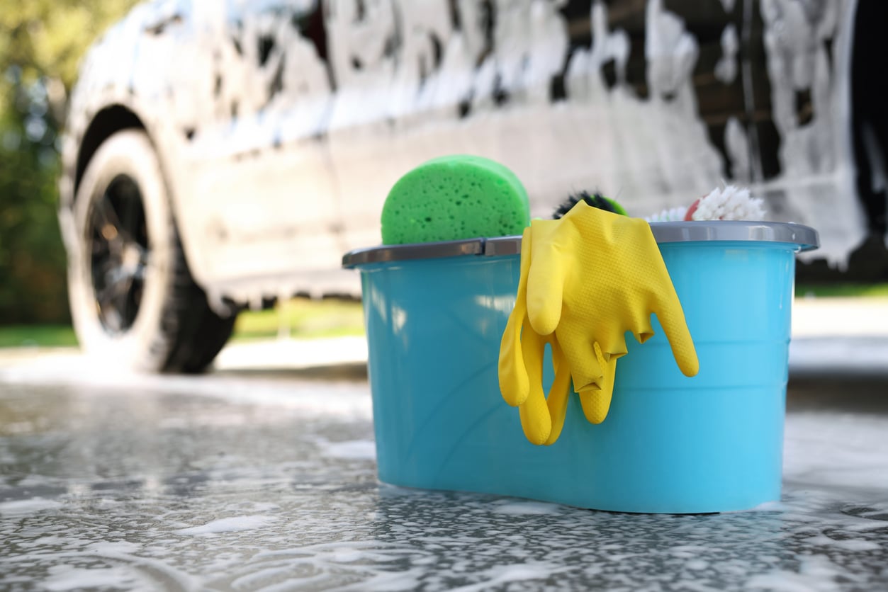 A blue bucket with a green sponge, yellow rubber gloves, and a brush sits on wet pavement near a soapy car being washed.