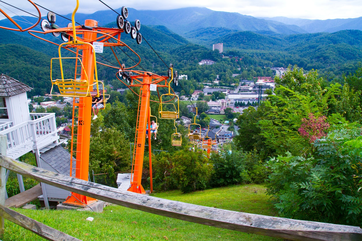Orange ski lift chairs ascend a grassy hill, overlooking a small town surrounded by lush green forests and distant blue mountains under a cloudy sky—a hidden gem among the travel spots boomers are learning to love.