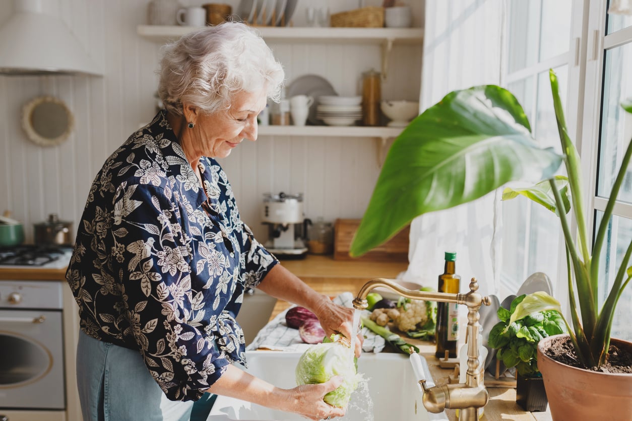 An older woman washes a head of cabbage at a kitchen sink, enjoying the simple pleasures that come with following some wise retirement tips. Sunlight streams in through a window, illuminating various vegetables and kitchen items on the wooden countertop. A large green plant sits by the window, adding to the tranquil, homey atmosphere.