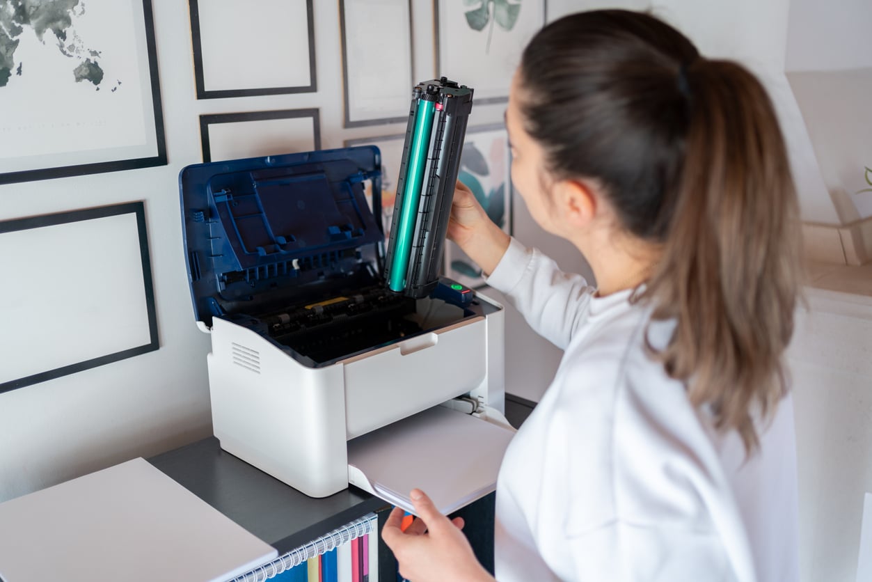 A woman with a ponytail replaces a toner cartridge in a white printer on a desk, with framed pictures on the wall in the background.
