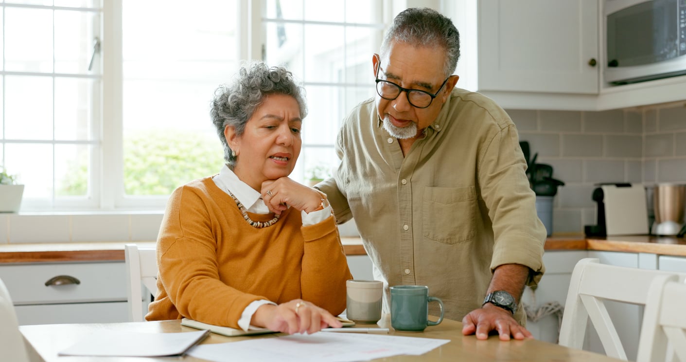 An older couple sits at a kitchen table, looking at papers together. The woman points at the documents while the man leans in to read, both appearing focused and thoughtful as they review retirement tips. Coffee mugs sit on the table beside them.