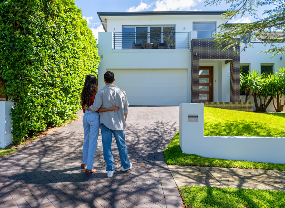 A couple stands arm in arm on a driveway, looking at a modern two-story house with large windows, a balcony, and a green lawn under a bright, sunny sky.