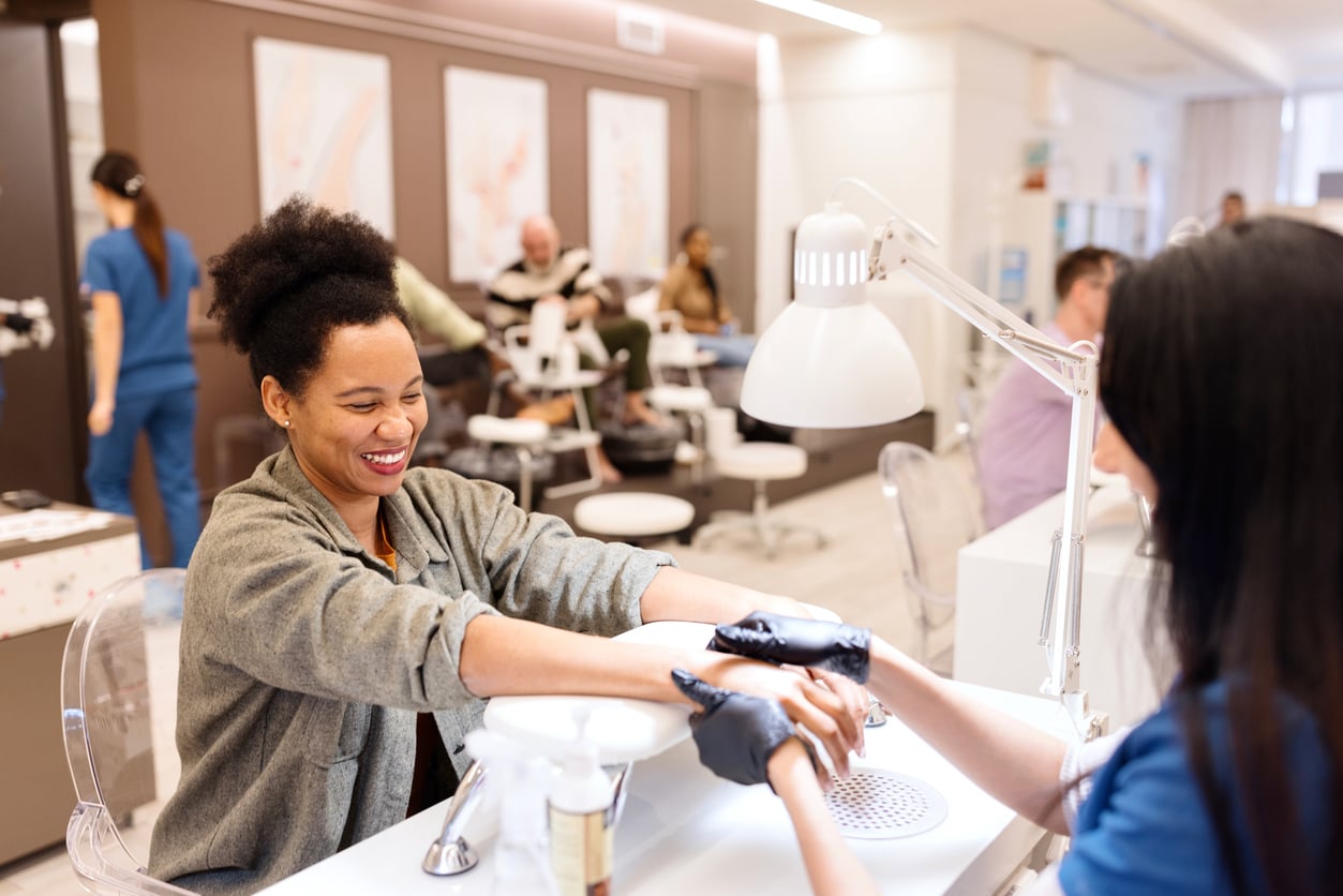 A woman smiles while receiving a manicure at a nail salon. The manicurist wears gloves. Other people are visible in the background, also getting nail or spa treatments. The atmosphere looks bright and welcoming.