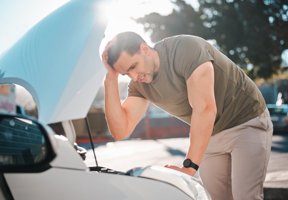 A man looks frustrated while examining the engine under the hood of his car on a sunny day.