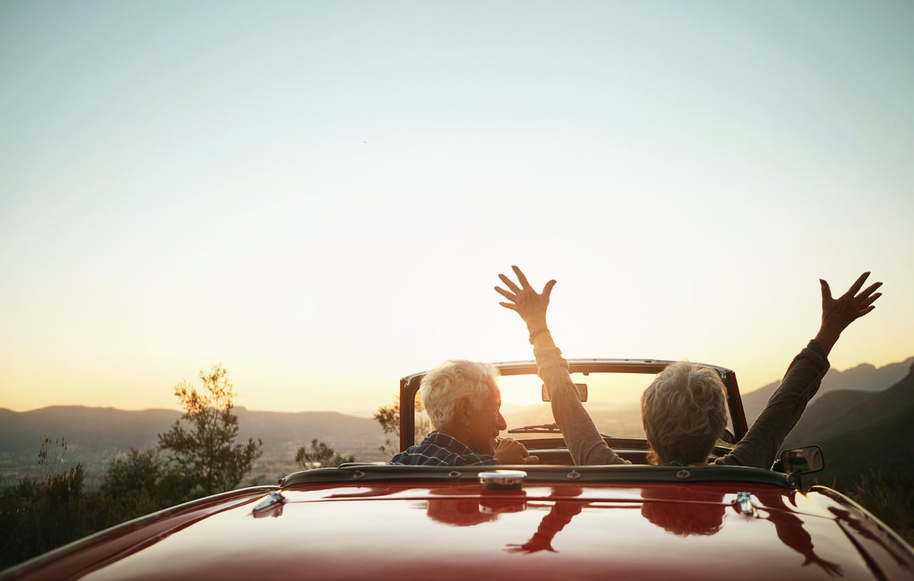 Two older adults ride in a red convertible at sunset; one person has both arms raised in joy. Their adventurous travel habits are evident as the car cruises through a scenic landscape, with hills and trees silhouetted against the sky.