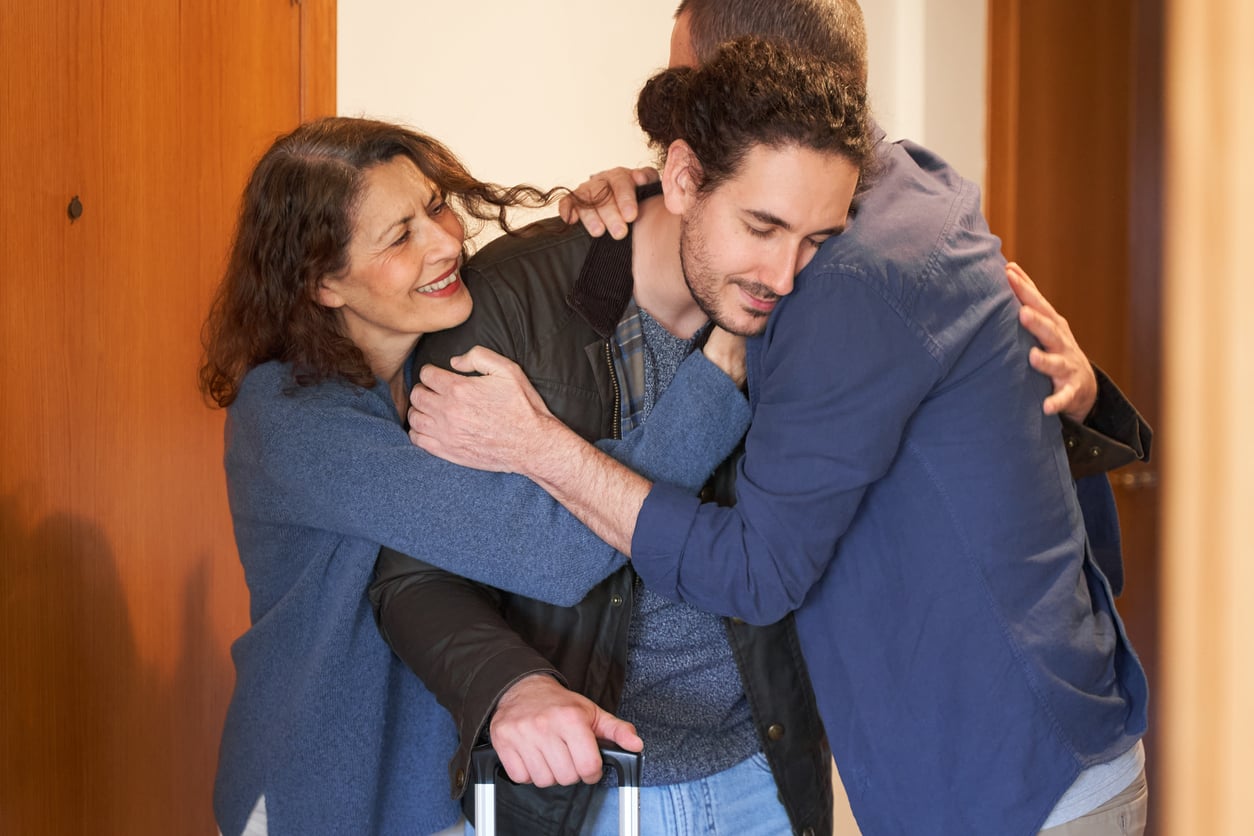 A young man with a suitcase hugs an older man and woman at a doorway. The woman smiles warmly, while the men embrace tightly, suggesting a heartfelt reunion.