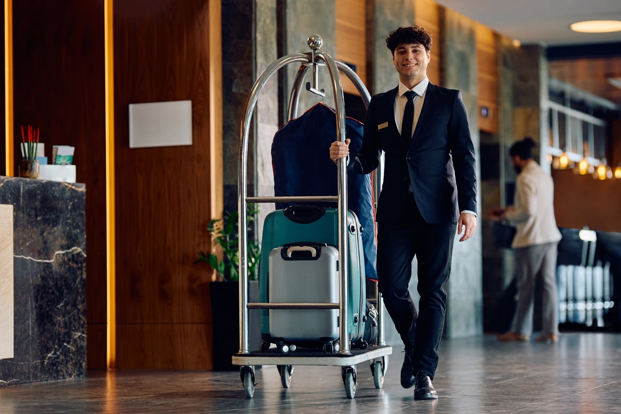 A hotel staff member in a suit smiles while pushing a luggage cart with suitcases in a modern hotel lobby, reminiscent of the personalized service and attention to detail among the things hotels used to offer guests. Another person stands in the background near the counter.