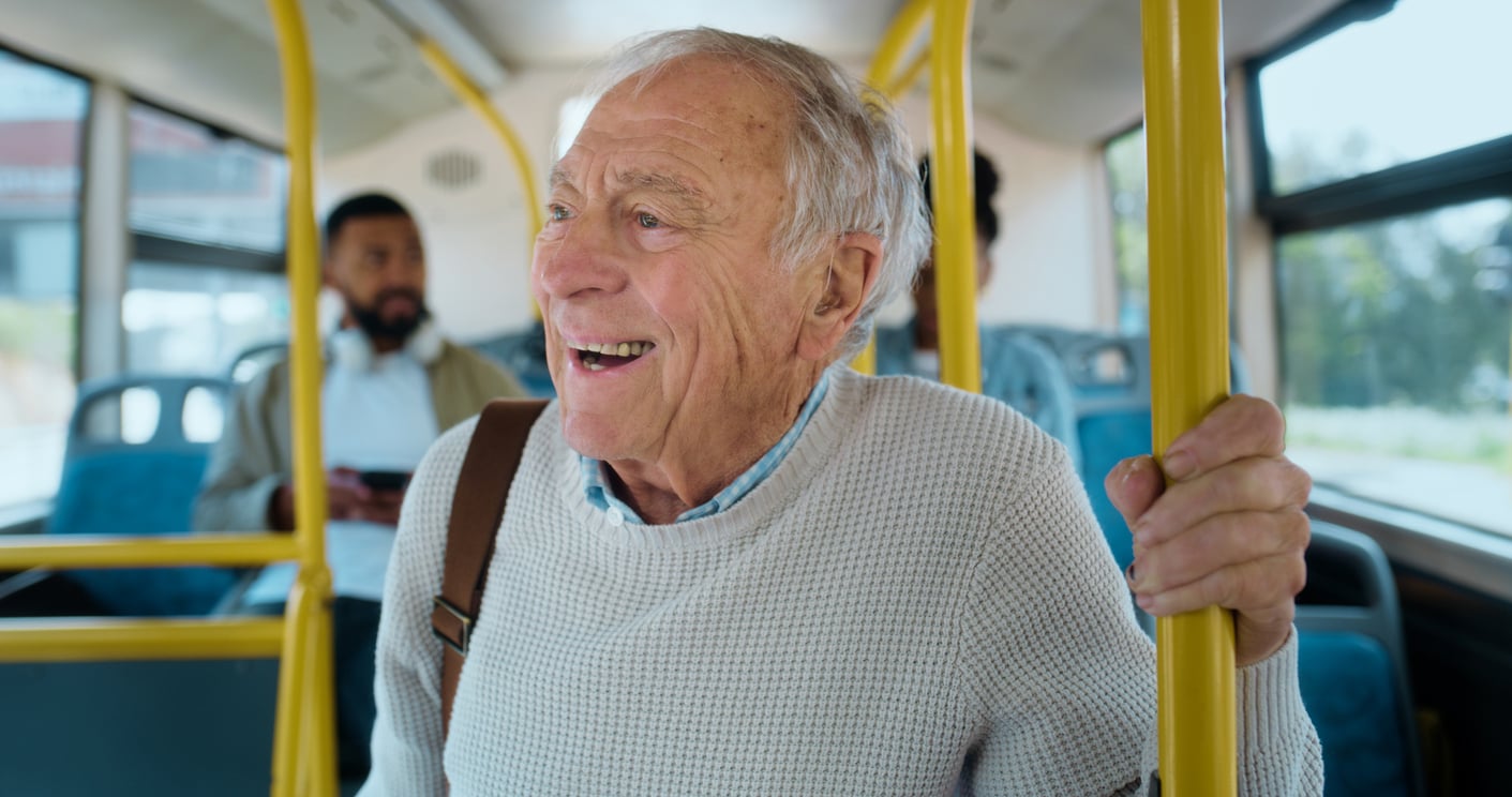 An older man with gray hair, wearing a light sweater and carrying a shoulder bag, smiles while standing and holding a yellow pole on a city bus. Other passengers are visible in the background.