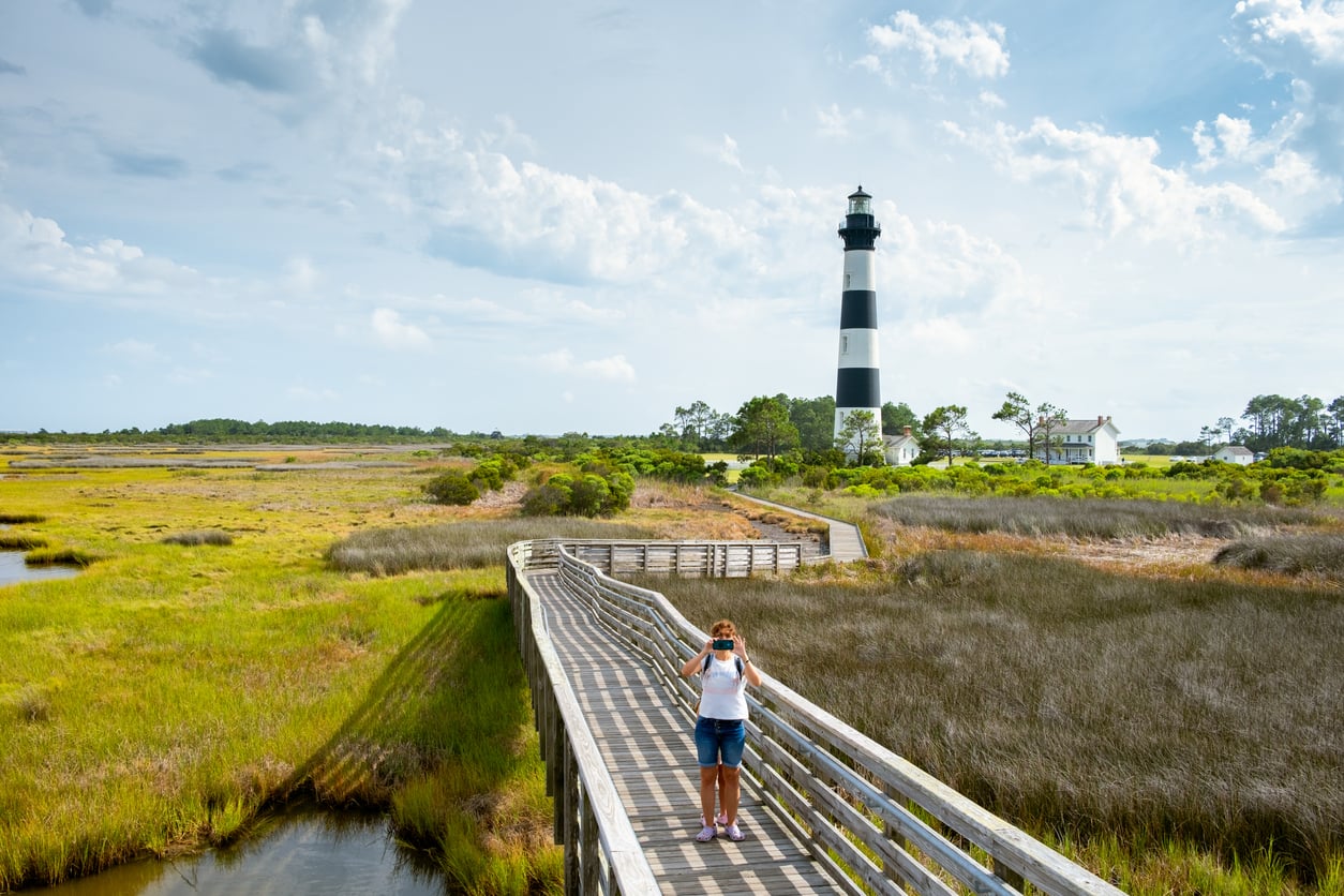 A person on a wooden boardwalk takes a photo in a grassy marsh with a black-and-white striped lighthouse and houses in the background under a partly cloudy sky, capturing one of the area’s most iconic vacation spots.