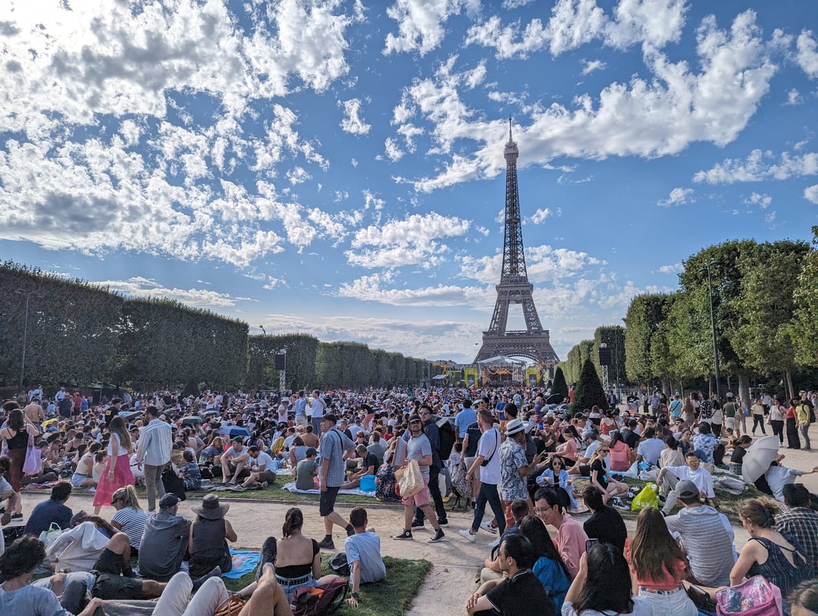 A large crowd of people sits and stands on the grass, with the Eiffel Tower in the background under a blue sky with scattered clouds.