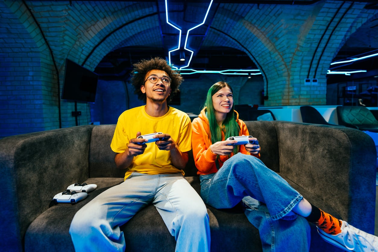 Two people sit on a gray couch playing video games with controllers. The person on the left wears a yellow shirt; the person on the right has green hair and an orange hoodie. The room has blue neon lights and brick walls.